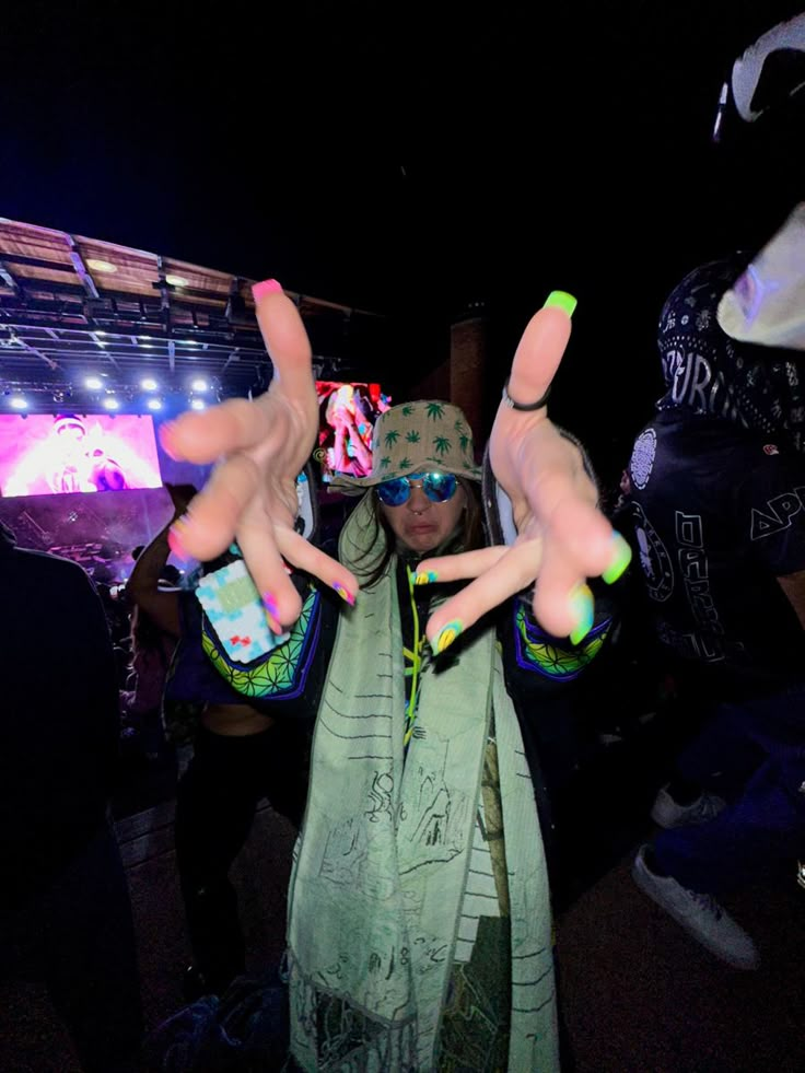 a woman with her hands up in the air at a music festival, wearing neon green and pink nail polish