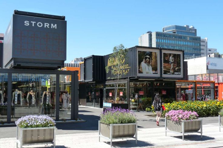 people are walking on the sidewalk in front of some buildings and flower pots with flowers