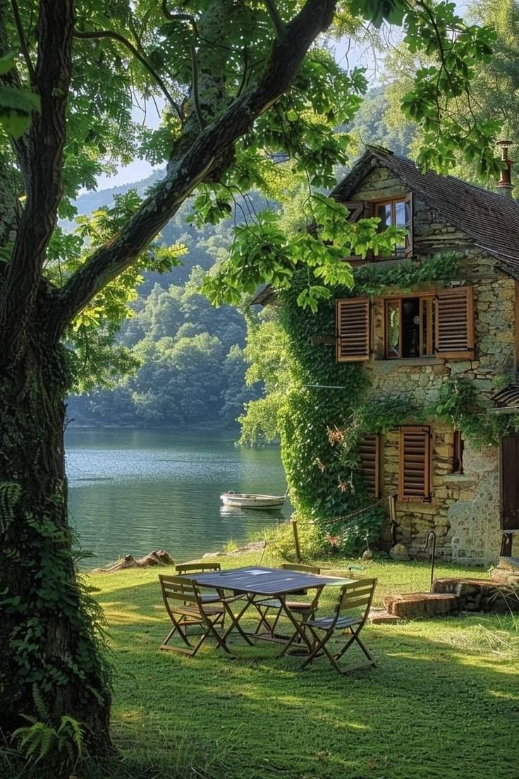 a table and chairs in front of a house by the water with trees around it