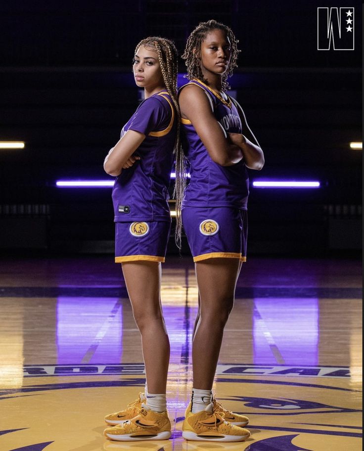 two female basketball players standing in front of a purple and yellow court with their hands on their hips