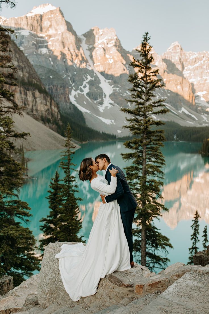 a bride and groom kissing on top of a mountain overlooking a lake with mountains in the background