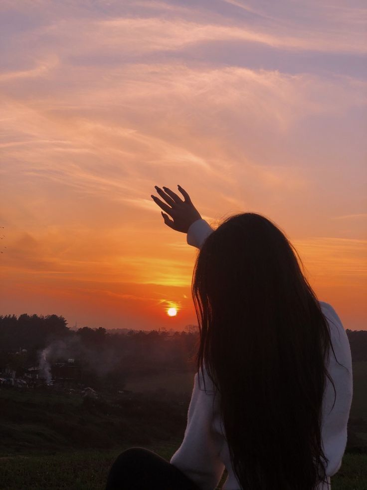 a woman sitting on top of a lush green field next to a sky filled with clouds