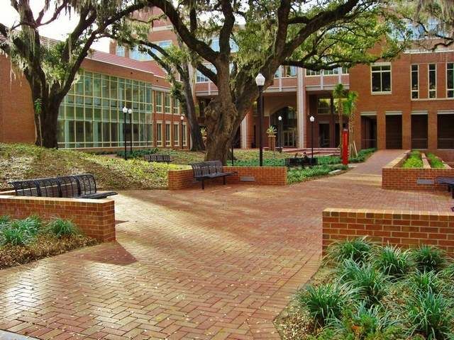 a brick courtyard with benches and trees in front of a building that has many windows