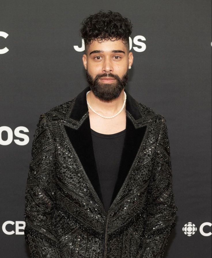 a man with a beard standing in front of a black background wearing a suit and pearls