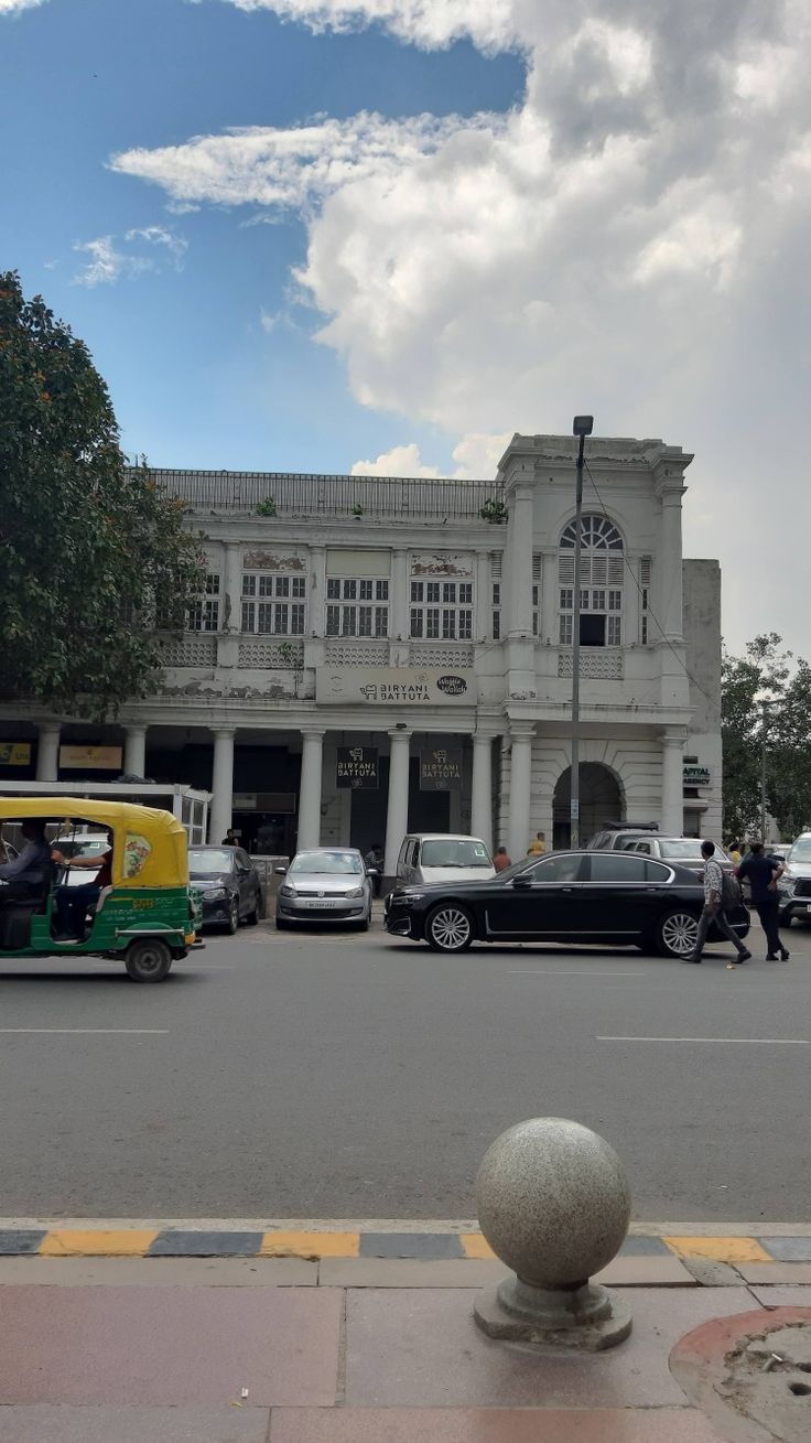 cars are parked in front of an old white building with columns on the side and people walking by