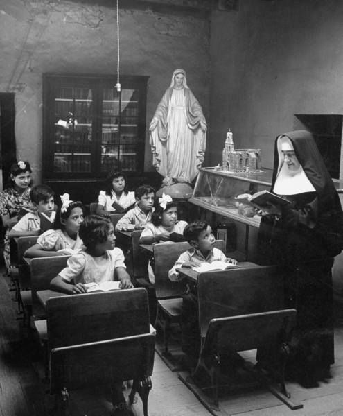 an old black and white photo of children sitting in school desks with the statue of jesus behind them