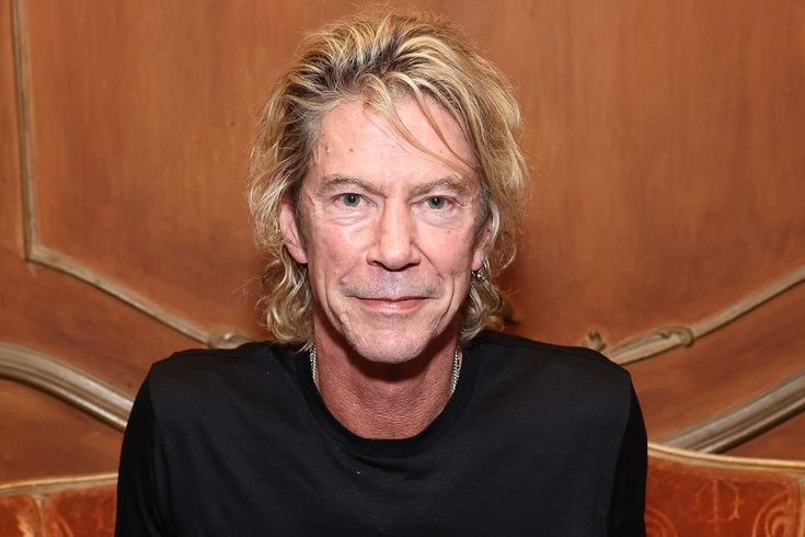 an older man with blonde hair sitting in front of a wooden paneled wall and looking at the camera