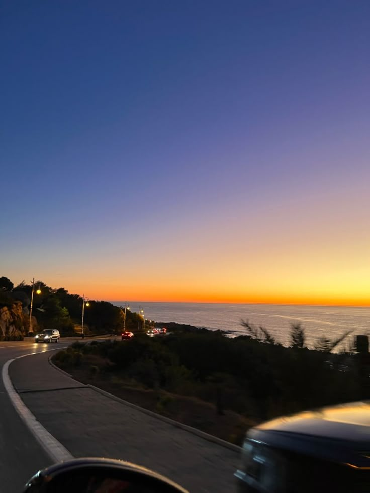 the sun is setting over the ocean and road with cars driving on it, as seen from inside a car