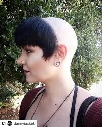 a woman with shaved hair and piercings standing in front of some bushes, looking off to the side