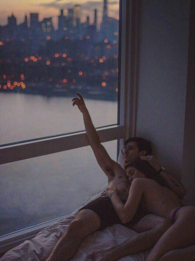 two people laying on a bed in front of a window looking out at the city