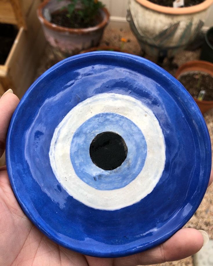 a hand holding a blue and white bowl with an eye painted on the side in front of potted plants