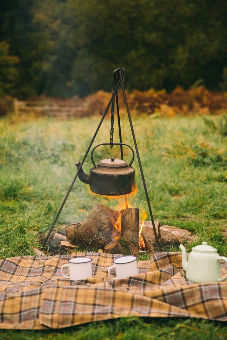 a campfire with two tea cups and a kettle on it next to a blanket