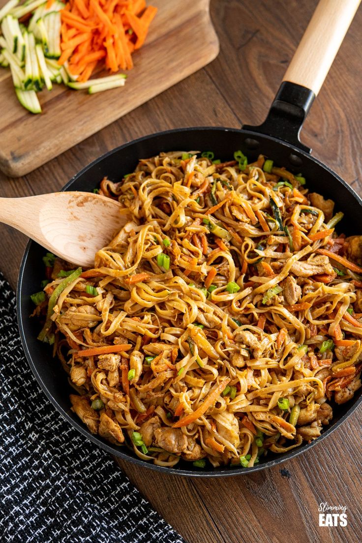 a pan filled with noodles and vegetables on top of a wooden table next to a cutting board