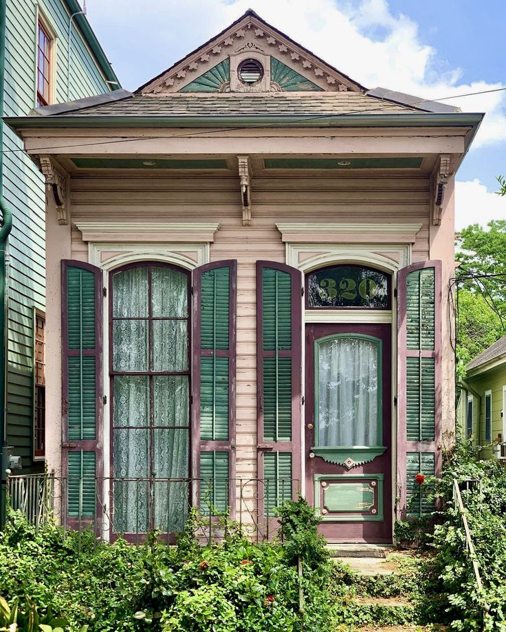 an old pink house with green shutters on the front and side windows, surrounded by greenery