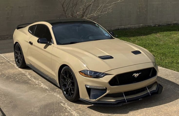 a beige mustang parked in front of a building with grass and bushes behind it on the sidewalk