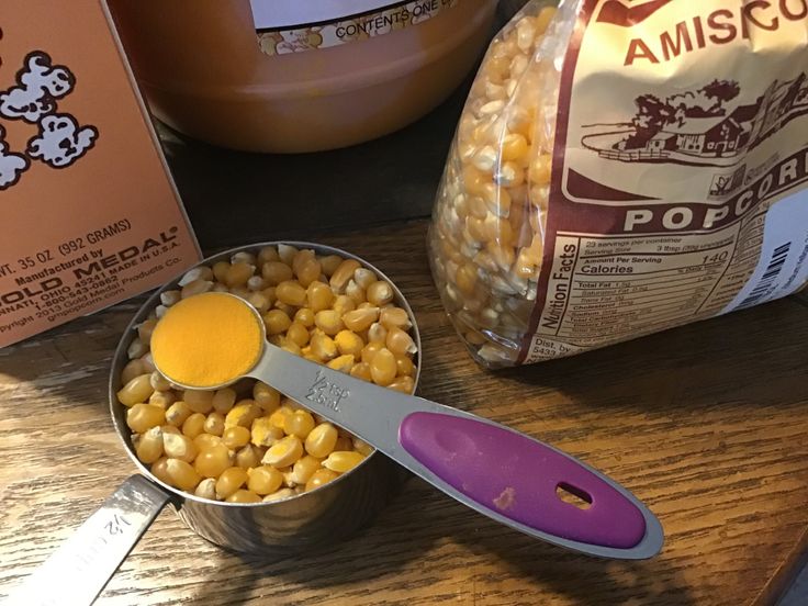 corn kernels are being spooned into a tin on a table next to two packages of popcorn