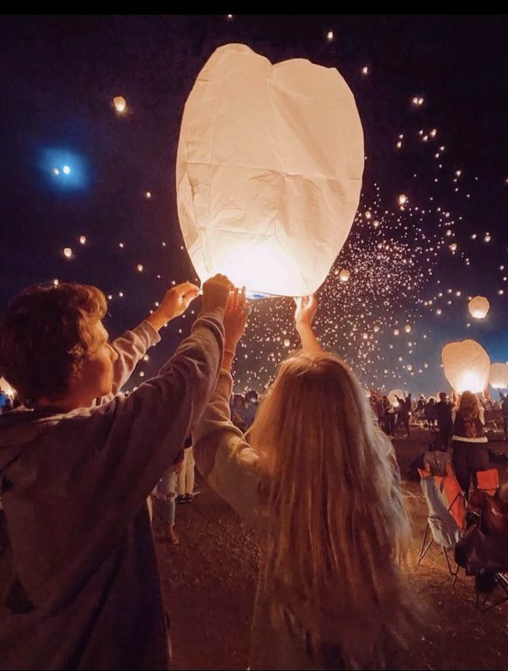 two people releasing paper lanterns into the sky at night with other people in the background