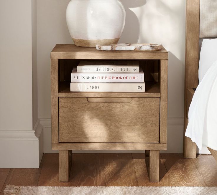 a nightstand with two books and a white vase on top of it next to a bed