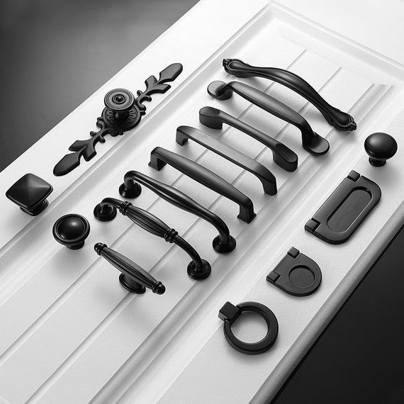 an assortment of black handles and knobs on a white cabinet door, all in different shapes and sizes