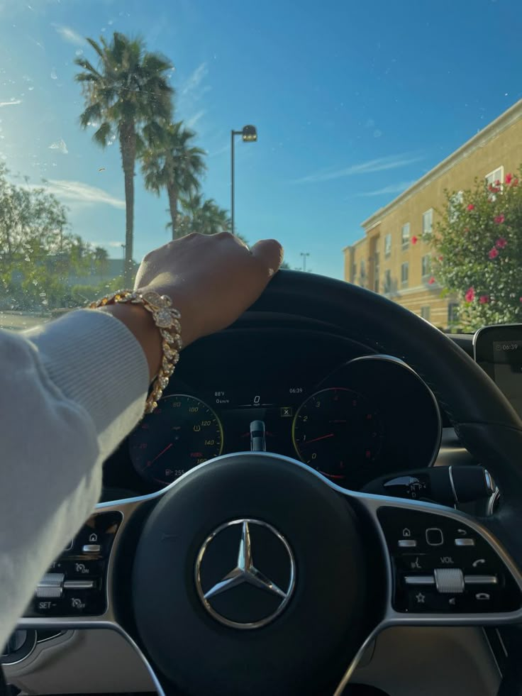 a person holding the steering wheel of a car with palm trees and buildings in the background