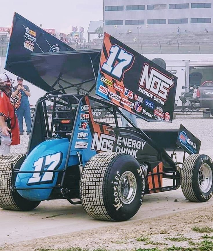 a man standing next to a blue and black race car on top of a dirt field