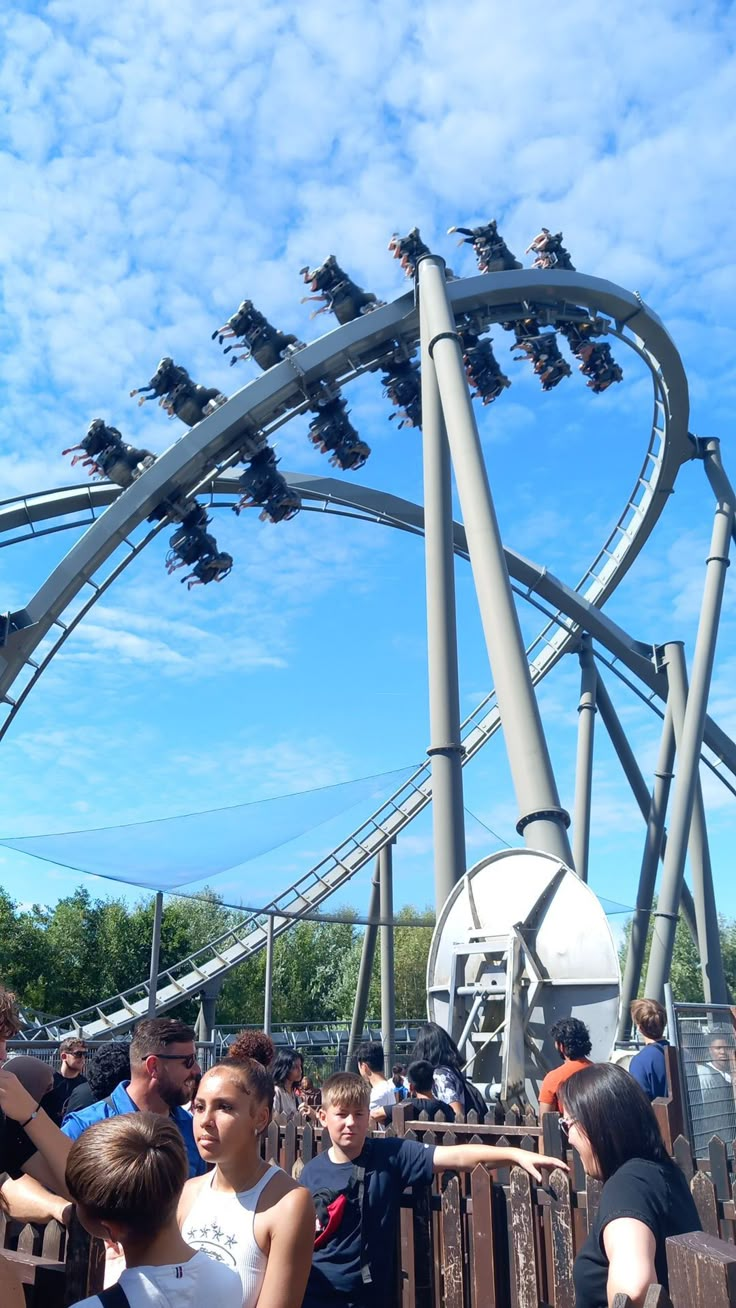 several people are standing in front of the roller coaster at an amusement park with blue skies and white clouds