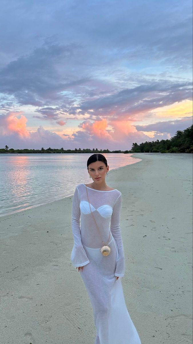 a woman standing on top of a sandy beach next to the ocean at sunset, wearing a white dress