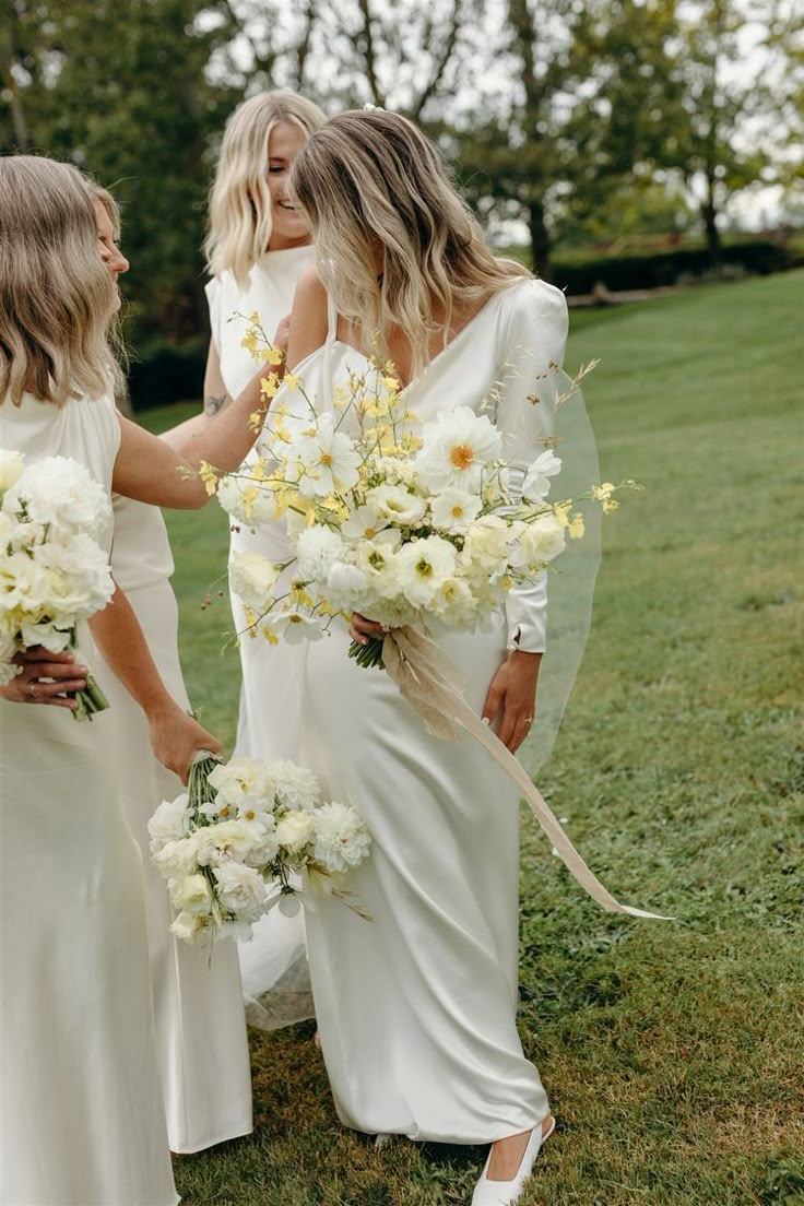 three bridesmaids in white dresses are holding bouquets and looking at each other