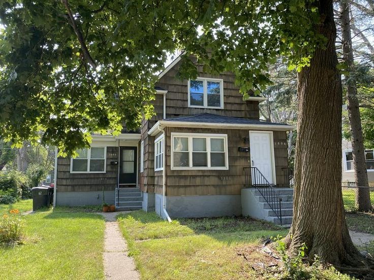 a small brown house sitting in the middle of a tree filled yard next to a sidewalk