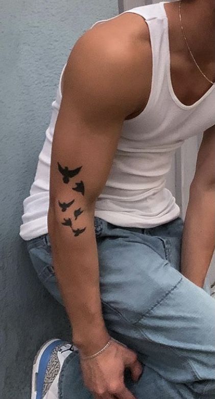 a man with a bird tattoo on his arm sitting against a wall and looking at the camera