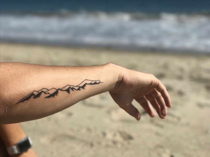 a person's arm with a mountain tattoo on it, in front of the ocean