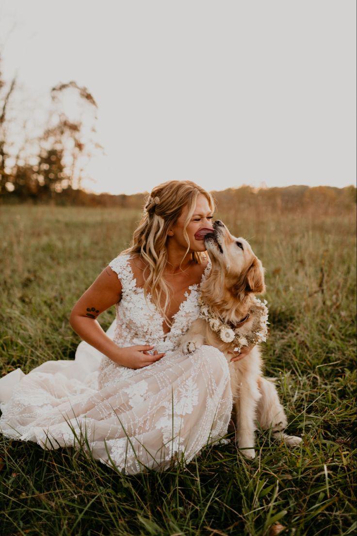 a woman in a wedding dress sitting on the grass with her dog and kissing her nose