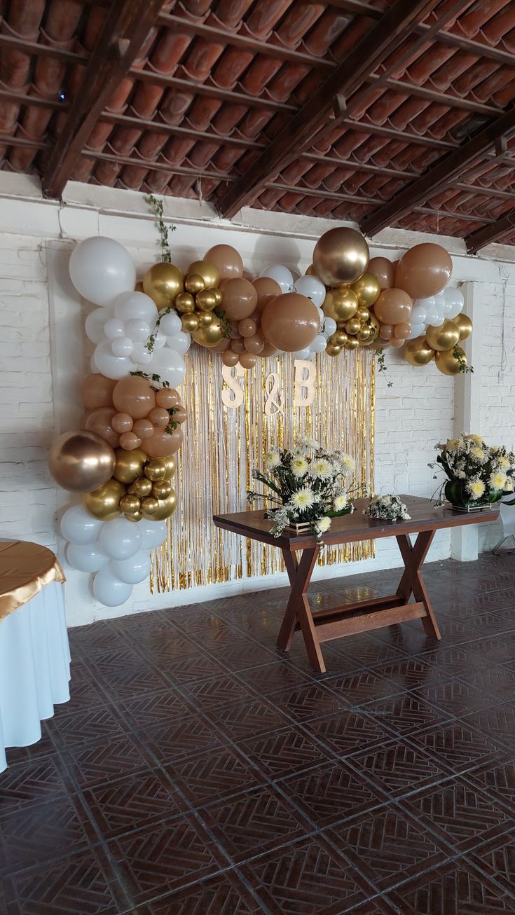 a table with flowers and balloons on it in front of a white brick wall that has gold foil streamers hanging from the ceiling