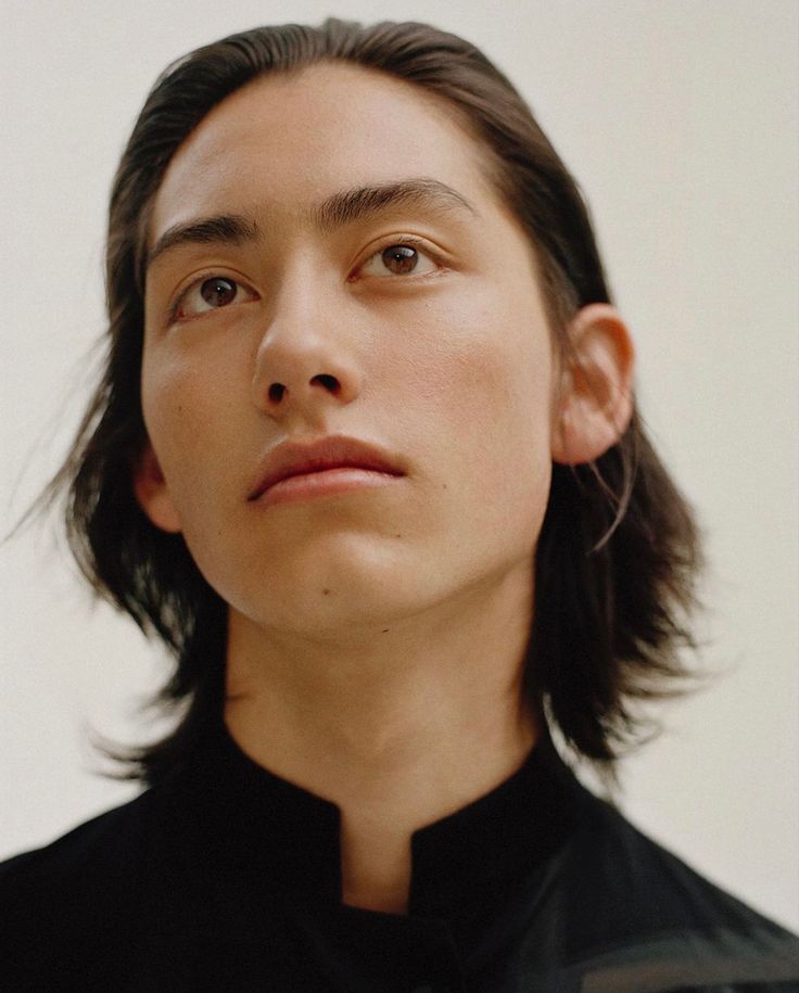 a man with long hair wearing a black shirt and looking off to the side while standing in front of a white wall