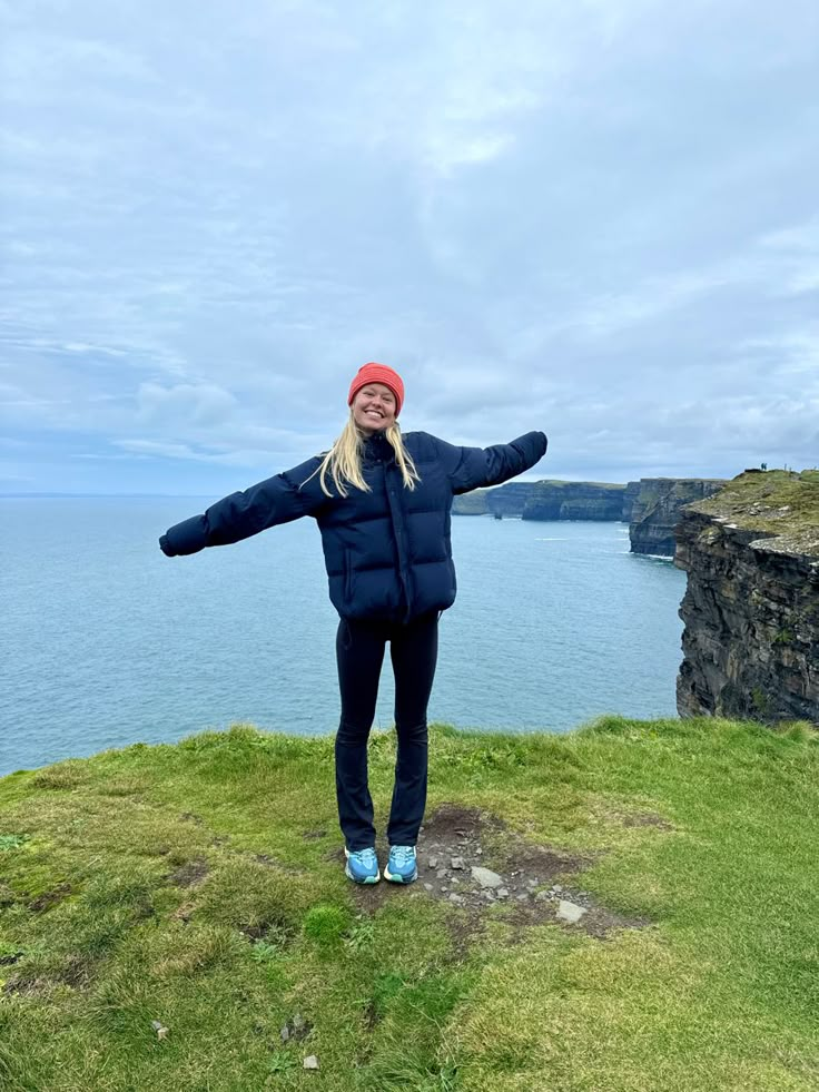 a woman standing on top of a grass covered hill next to the ocean with her arms outstretched