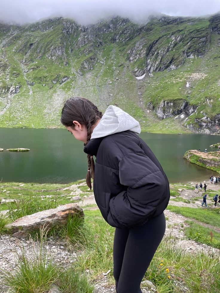 a woman standing on top of a grass covered hillside next to a lake with mountains in the background