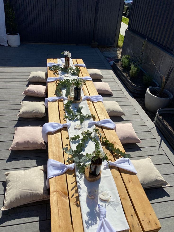 an outdoor table set up with white linens and greenery on it, along with pillows