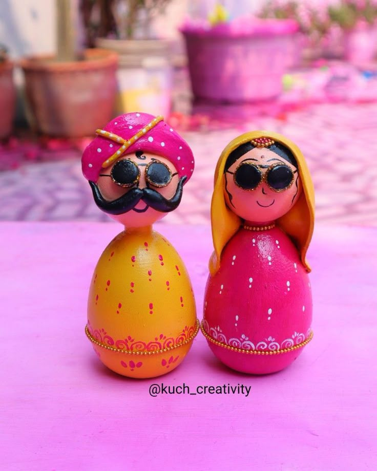 two wooden dolls sitting next to each other on a pink tableclothed surface with potted plants in the background