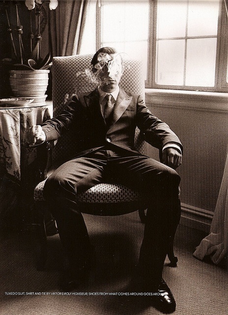 black and white photograph of man in suit sitting in chair next to large window with potted plant