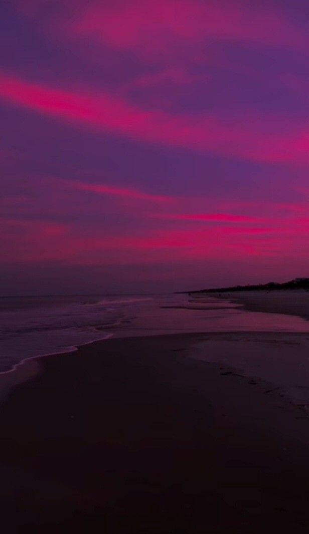 the sky is pink and purple as it sets over the water at dusk on the beach