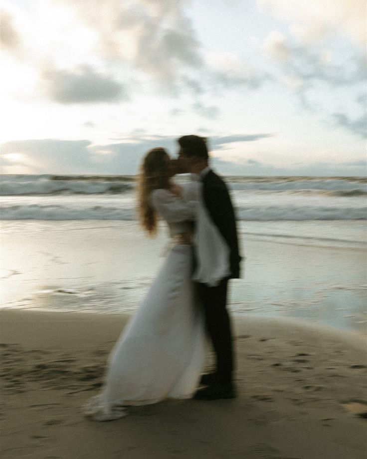 a bride and groom standing on the beach at sunset in front of the ocean with their arms around each other