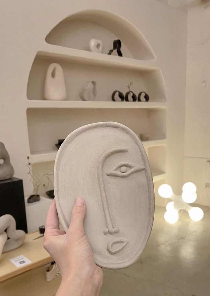 a person holding up a clay mask in front of a shelf