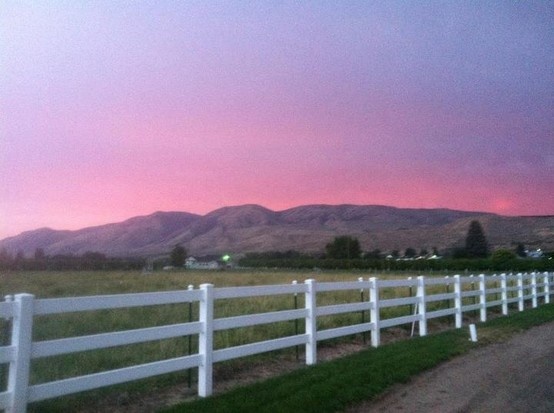 a white picket fence in front of a field with mountains in the background at sunset