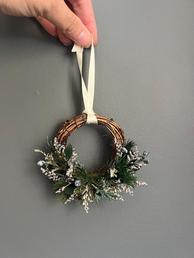 a hand holding a christmas wreath hanging from the side of a wall with white ribbon