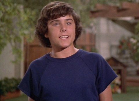 a young boy standing in front of a house wearing a blue t - shirt and smiling