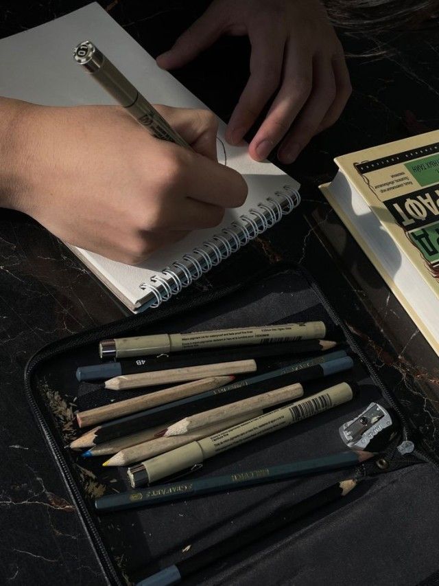 a person is writing on a notebook next to some pencils and an open book