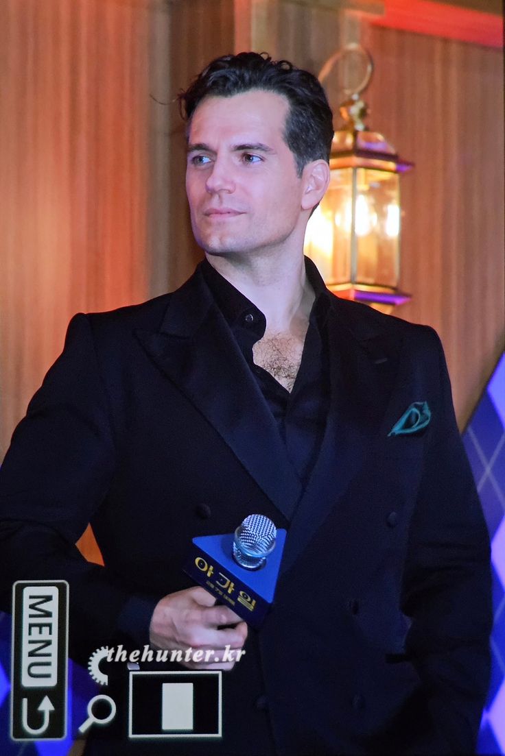 a man in a black suit holding a microphone and looking at the camera while standing next to a wall