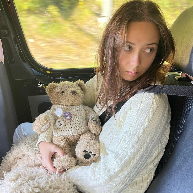 a woman sitting in the back seat of a car holding a teddy bear and wearing a sweater