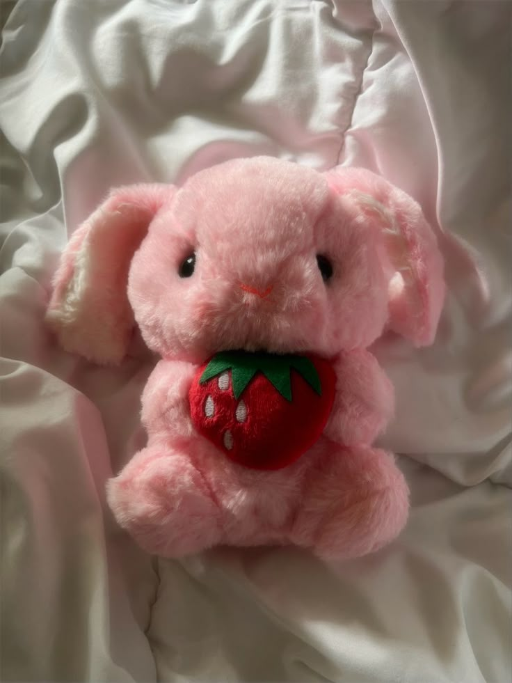 a pink stuffed animal with a strawberry on it's chest sitting on a white sheet