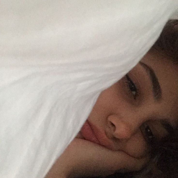 a woman laying in bed under a white blanket with her head on the pillow and looking at the camera